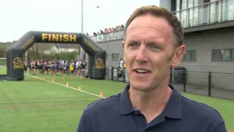 Grant Rees wearing a dark polo top and smiling with the finish arch behind him on the green grass of the pitch.