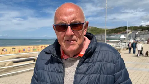 A bald man with a blue puffer jacket looking at the camera. He has sunglasses on and is stood on the promenade.