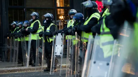 Ian Forsyth/Getty Images A line of police officers in riot gear. They hold transparent shields in front of them.