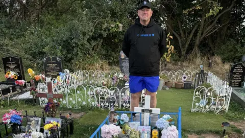 Family A man wearing a black hoodie and blue shorts stands behind a gravesite. The hoodie has the words 'Forever 23' written on the front.
