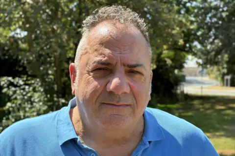A middle-aged man with short cropped hair, curly on top, stands in sunlight in front of a patch of grass and green trees. He is wearing a blue polo shirt and has a serious expression on his face.