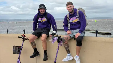 Supplied Two men in purple hoodies, wearing shorts and trainers and backpacks, are sitting on a promenade in front of the sea with two purple scooters in front of them.