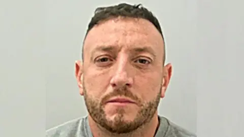 Police custody image of Jake Balmforth looking straight at the camera. He has short brown hair and stubble and is wearing a grey top.
