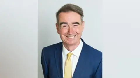 Wokingham Borough Council Stephen Conway is wearing a blue suit jacket, a yellow tie and a white shirt. He is smiling and has short grey hair. He is standing in front of a grey background.