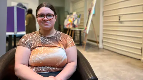 Elianna looks at the camera as she sits in a chair. Behind her is a corridoor with an art piece in the middle of the area. Elianna is wearing a light brown patterned t-shirt and her brown her is tied into a ponytail. 