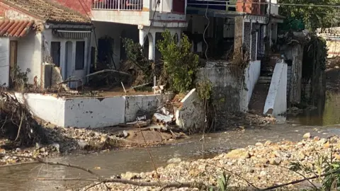 Diana Whitwell Water flows past damaged homes on Spanish mountain. Some of the garden walls have collapsed, there is rubble and vegetation around.