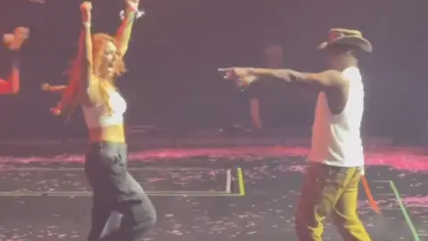 A woman with long red hair, a white tank top and black cargo trousers stands on a black stage stage with her arms in the air. To the right singer Ne-Yo, wearing a white vest top, mustard coloured trousers and a brown cowboy-style hat points towards her with his right hand. 