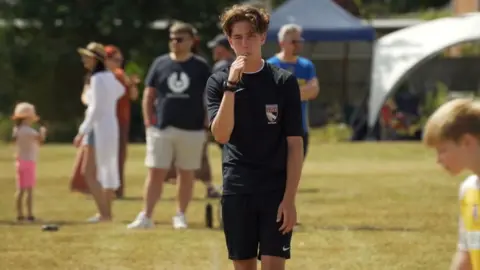 Jonny Michel/BBC Alex blowing the whistle during a football match. He has brown hair and is wearing his referee uniform. A number of spectators are behind him.