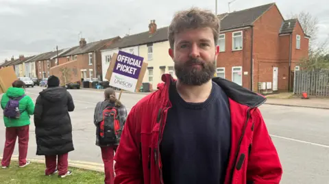 Christopher Roche stands on a roadside grass verge as three women behind him hold signs on a picket line. There are terraced houses on the other side of the road. Mr Roche has a beard and is wearing a red coat and a navy jumper.