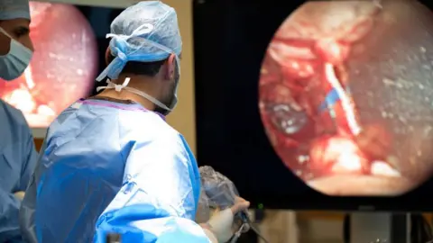 PA Media The image shows a surgical procedure taking place in an operating theatre. The focus is on a large monitor displaying a magnified internal view of a brain. In front of the monitor, a surgeon dressed in blue surgical scrubs is holding a medical instrument. 