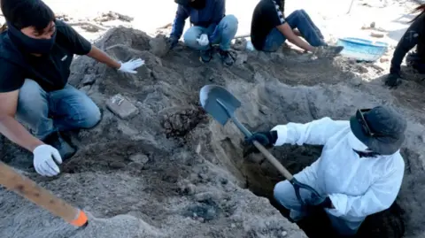 Man in white shirt is holding a trowel as he stands in a hole in the ground