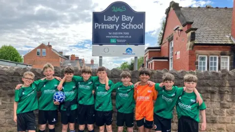 9 boys in school football team stood in front of Lady Bay Primary School wall in green football kit.