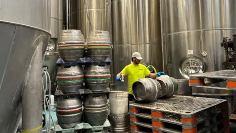 Charlie Stubbs/BBC There is a man in a yellow t-shirt and white cap working on two kegs, which are on top of a black board. This is supported by multiple layers and orange bands supporting each leg. There are six kegs to his left and five brewing vats around him