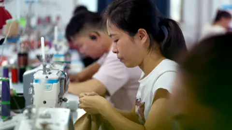 A woman uses a  sewing machine with other factory workers in view, making an outfit in a factory that supplies to Shein.