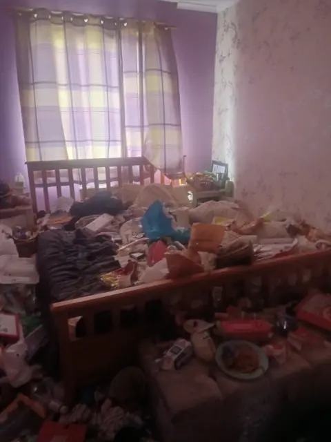 A bedroom with the curtains shut but light trying to escape through and a wooden double bed covered in rubbish.