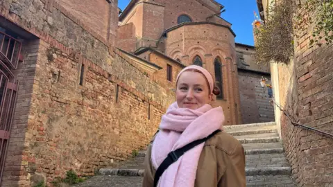 Nell Eustace Nell Eustace, a student from Bristol currently studying in Italy, is pictured in Italy. She is wearing a pink scarf and headband, and is standing before an ancient building and stone stairs.