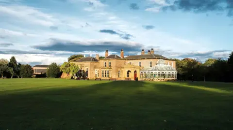 Preston Hall, a large manor house with an attached atrium, sitting at the centre of green lawns with trees visible in the distance