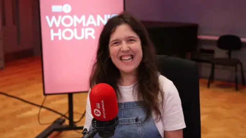 Actor and comedian Rosie Jones smiles in the Woman's Hour studio - she has long dark hair and wears denim dungarees