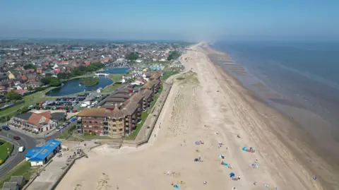 A aerial view of a coastline on a sunny day showing the sea, a broad beach and homes and businesses inland under a blue sky. 