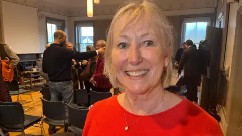 Frome resident Bridget Clark is standing in a room in a red shirt. She is smiling at the camera. Behind her are chairs and people talking in groups.