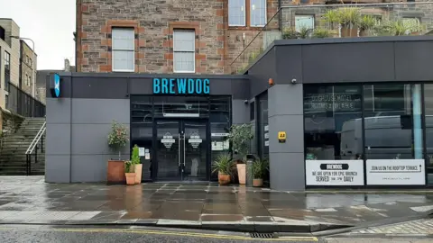 A Brewdog branded pub with blue and grey modern frontage on an old red stone building in the Old Town of Edinburgh. It has been raining and the pavement is wet.