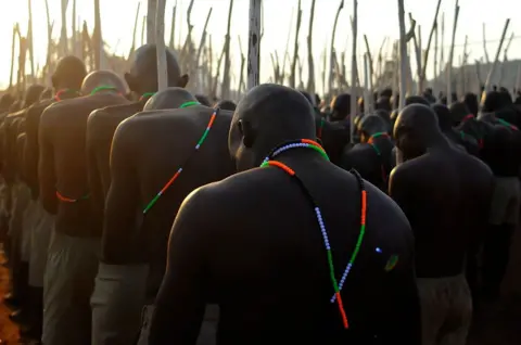 LUCAS LEDWABA / AFP / GETTY IMAGES Men wearing matching khaki trousers and coloured beads around their necks stand in formation with batons. Behind them, the sun is setting. The earth is a rich red colour.