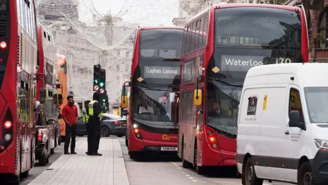A busy central London street with multiple red double-decker buses stuck in slow-moving traffic. A white van and several cars are also queued, while two men stand on the pavement beside a bus. Christmas lights hang above the road, and buildings line the background.