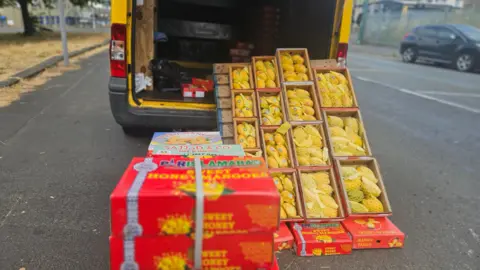 Aisha Iqbal/BBC A yellow van parked on the side of a road. Piled up outside it, and visible inside, are multiple small crates of bright, ripe mangoes. The fruits are strapped together in closed boxes, or are on display in open boxes with a soft protective netting covering them.