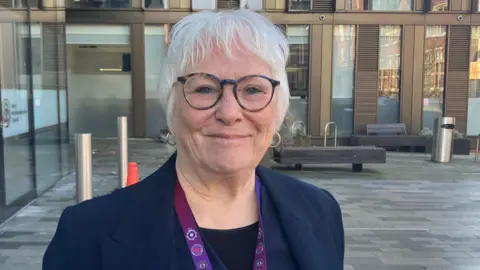 Local Democracy Reporting Service Danielle Stone is standing outside a modern glass-fronted building with block paving. She is standing and smiling while wearing a blue blazer, a purple police lanyard and tortoiseshell patterned eye glasses.