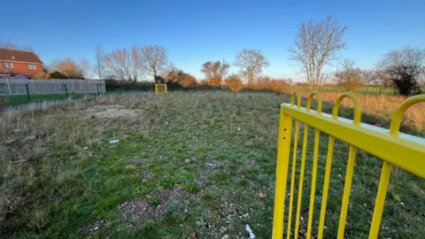 A view of an empty play area with overgrown grass and litter on the ground.