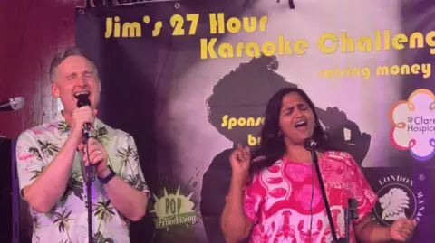 Toby Acton A man and a woman in loud, colourful tops singing with their eyes closed and mic stands in front of them. "Jim's 27 hour karaoke challenge is written in the background
