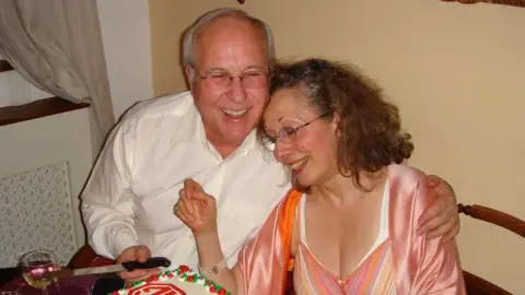 Rivka Gottlieb Michael Gottlieb is pictures hugging his wife Milli and cutting a cake on their 40th wedding anniversary. He is wearing a white shirt and is laughing. She is wearing a pink dress and is burying her head in his shoulder.