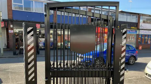 A black steel gate on a commercial street. Cars and shops are in the background. A plain black sheet of metal that should bear a coat of arms sits in the middle of the gate.