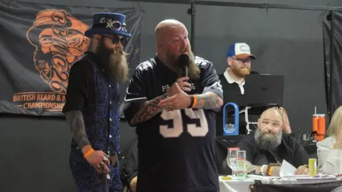 Shane Hazelgrave Lewis Aylmer wearing a black, long-sleeved shirt, a blue and black waistcoat, blue and black patterned trousers, black glasses and a blue top hat. He has a long, bushy, brown beard and a long, brown moustache. He is stood next to a man who is speaking into a microphone. He is wearing a blue sports top with numbers on the front. There are people sat behind a table in the background. There is also a black and orange banner hung on a black wall.