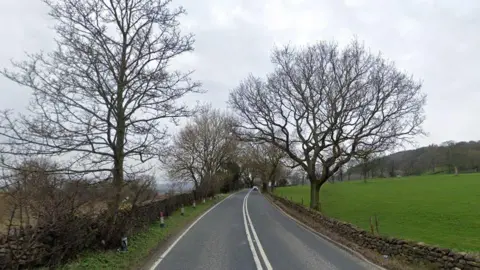 A rural A-road lined with trees, a stone wall and a field.