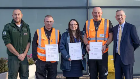 Five people standing next to each other in a line outside, looking at the camera. Three of them are holding certificates and wearing orange high-vis jackets.