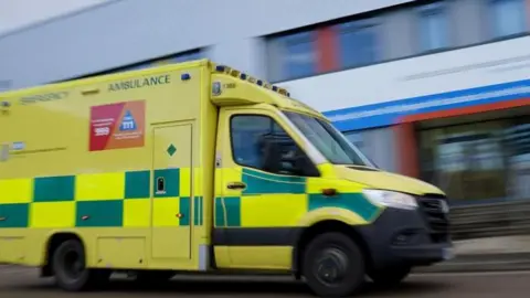 PA Media A photo of a green and yellow ambulance driving along a road, there is a blurred hospital in the background