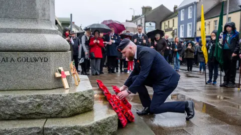 Dai Evans Photograph of a man kneeling on one knee to lay a poppy wreath. 