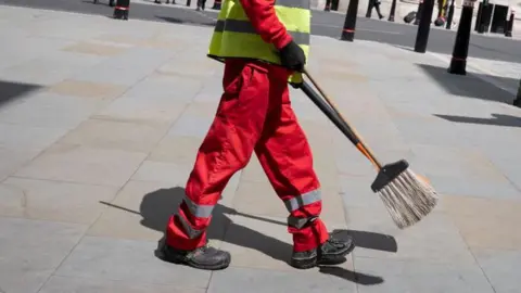 Getty Images A typical image of a street cleaner in London, wearing a red and white uniform and carrying a broom.