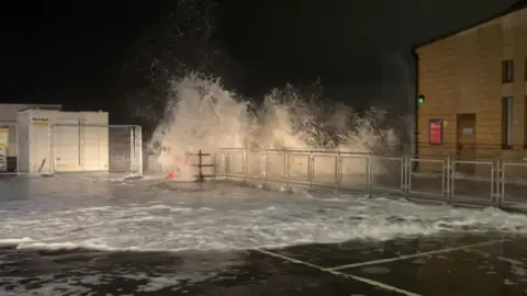 A large wave crashes over a metal barrier onto a waterfront walkway at night, lit by nearby buildings.