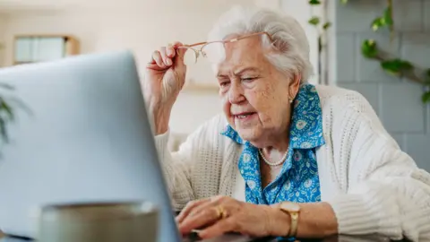Getty Images A woman with grey hair, blue blouse and white cardigan lifting up her glasses and looking at a computer monitor