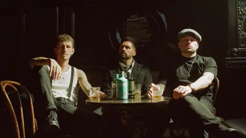 Jack Geddes From left to right shows Nathan, Stevie and Alan sitting at a table in a bar drinking whiskey, all dressed in early 1900s aesthetic attire.