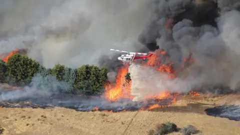 A helicopter drops water over a bushfire. Large plumes of dark smoke rise from the ground.