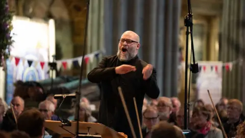 Michael Whitefoot A man with glasses and a grey beard is standing up with his mouth open and arms raised. Other people are sitting around him.