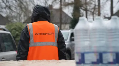 PA Media A person in an orange jacket. Behind them are some water bottles. 