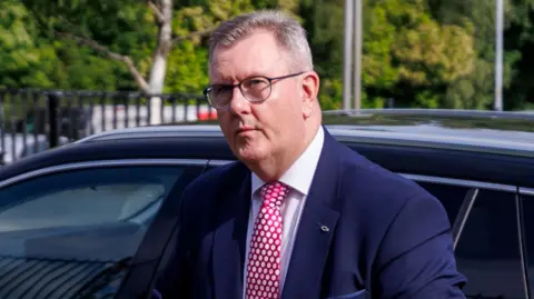 A man with short, greying hair is exiting a black car. He is wearing a blue suit jacket, white shirt and red tie with white polkadots. He has black-rimmed glasses on. In the background more parked cars, a metal fence, lampposts and trees are visible. 