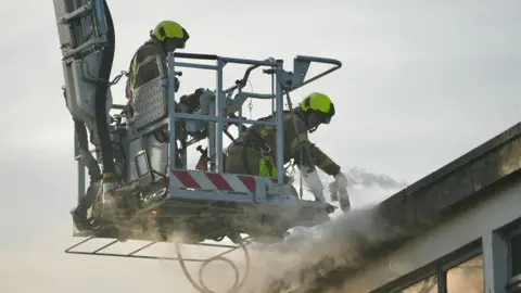 Martin Giles/BBC Two firefighters on an aerial ladder platform putting out the fire. There is smoke coming out of the top of the building.