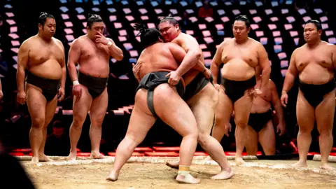 PA Media A line of four sumo wrestlers in the traditional black loincloth worn for the sport look on as two wrestlers grapple in front of them