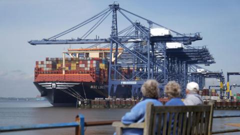 Three older people look towards the Port of Felixstowe while sitting on a bench, their backs turned