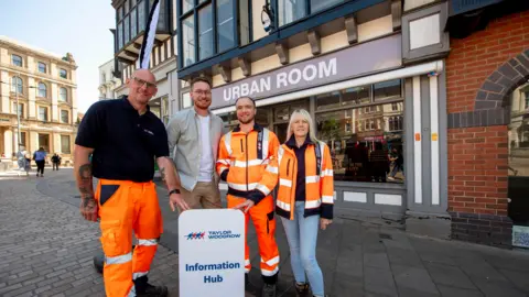 A woman and three men stand together outside the Urban Room in Wolverhampton city centre. Two of them touch a white sign saying 'Taylor Woodrow Information Hub' on it. The woman wears an orange hi-vis jacket and two of the men also wear hi-vis clothing. The other man is in a light coloured shirt with a white T-shirt underneath and brown trousers. 
